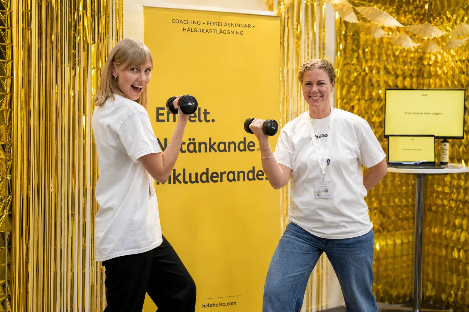 Två Helo-medarbetare lyfter hantlar tillsammans i en guldfärgad eventmonter
